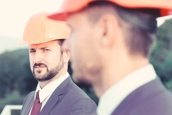 Los constructores controlan las obras. Concepto de construcción. Los gerentes usan trajes inteligentes, corbatas y sombreros en el fondo de la naturaleza, desenfocados. Líderes con barba y caras concentradas discuten proyecto — Foto de Stock