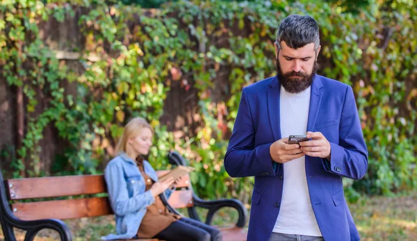 Hombre con barba mantenga el teléfono inteligente, mientras que la niña sentarse parque banco. Aplicación de uso del hombre para citas. Un tipo buscando novia. Concepto de aplicación de citas. Solicitud de búsqueda pareja relaciones románticas —  Fotos de Stock