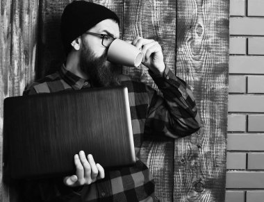 Sakallı, beyaz tenli hippi elinde bardakla dizüstü bilgisayar tutuyor.