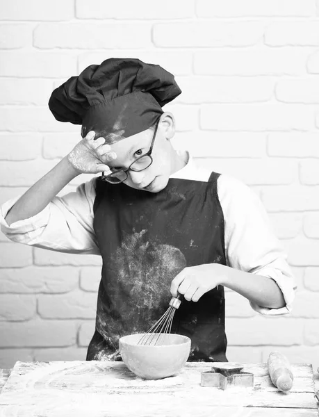 Kleiner Junge netter Koch Koch in roter Uniform und Hut auf gefärbtem Gesicht Mehl mit Gläsern am Tisch stehend mit Nudelholz und Kochen mit Küchenbesen in türkisfarbener Schüssel auf weißem Backsteinmauerhintergrund — Stockfoto