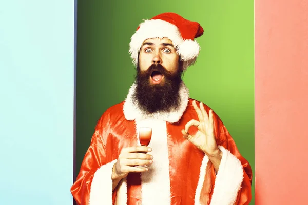 Knappe man van de baard Kerstman met lange baard op verbaasd gezicht houden glas alcoholische schot in rode kerst of xmas vacht en Nieuwjaar hoed op kleurrijke studio achtergrond — Stockfoto