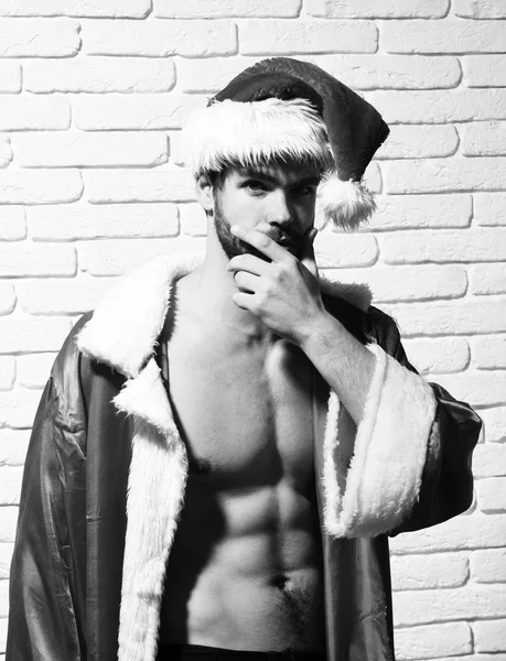 Giovane bell'uomo di Natale sexy barbuto con elegante barba in cappello rosso Babbo Natale e cappotto con busto nudo muscolare su sfondo muro di mattoni bianchi — Foto Stock