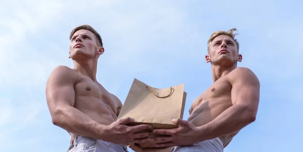 Hot sales and discount. Brothers buy eco healthy products. Guys attractive twins carry shopping bag made out of brown paper. Sales season. Men muscular athletes hold shopping bag sky background