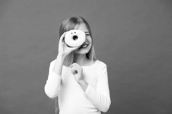 Chica en la cara sonriente sostiene donut dulce en la mano, fondo violeta. Niña con el pelo largo le gustan los dulces y golosinas. Concepto de diente dulce. Chica mirando a través del agujero en donut con un par de ojos — Foto de Stock