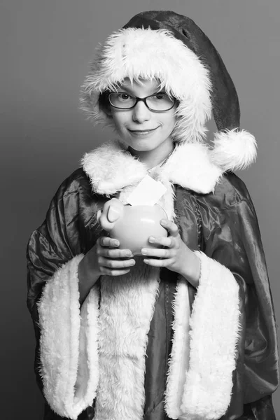 Młody chłopak ładny Święty Mikołaj z okulary w sweter Boże Narodzenie i nowy rok christmas hat przytrzymanie różowy świnia piggy bank na tle czerwony studio — Zdjęcie stockowe