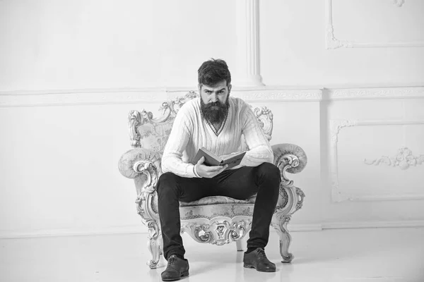 Homme à la barbe et à la moustache assis sur un fauteuil et un livre de lecture, fond mural blanc. Scientifique, professeur au visage sérieux explore la littérature. Macho smart passe ses loisirs avec le livre. Concept de connaissance — Photo