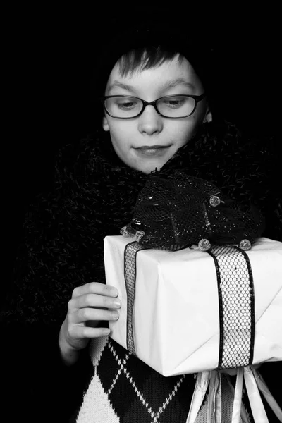 Pequeno menino sorridente ou garoto nerd bonito em óculos, chapéu e cachecol de malha na moda no fundo preto mantém caixa de presente branco com bo vermelho — Fotografia de Stock
