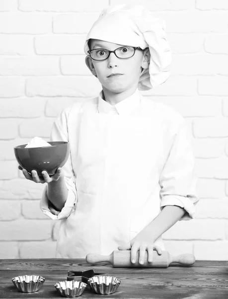 Jeune garçon petit chef cuisinier mignon en uniforme blanc et chapeau sur le visage surpris avec des lunettes debout près de la table avec des moules à rouleau pour gâteaux et tenant bol rouge sur fond de mur de brique — Photo