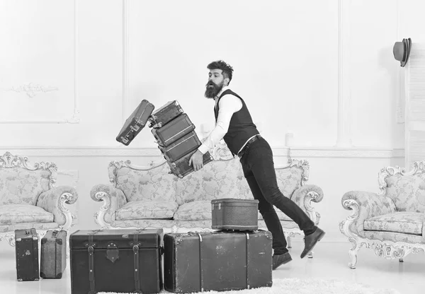 Porter, butler accidentally stumbled, dropping pile of vintage suitcases. Baggage insurance concept. Man with beard and mustache in classic suit delivers luggage, luxury white interior background