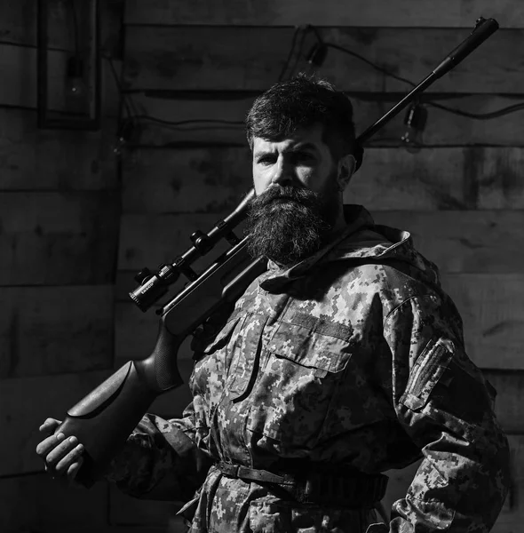 Hunter, brutal hipster on strict face with gun ready for hunting. Gamekeeper concept. Man with beard wears camouflage clothing, carries rifle on shoulder, wooden interior background — Stock Photo, Image