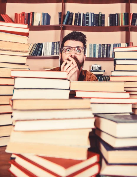 Homme au visage choqué entre des piles de livres à la bibliothèque, étagères sur fond. Concept de découverte scientifique. Enseignant ou élève barbu porte des lunettes, s'assoit à table avec des livres, déconcentré — Photo