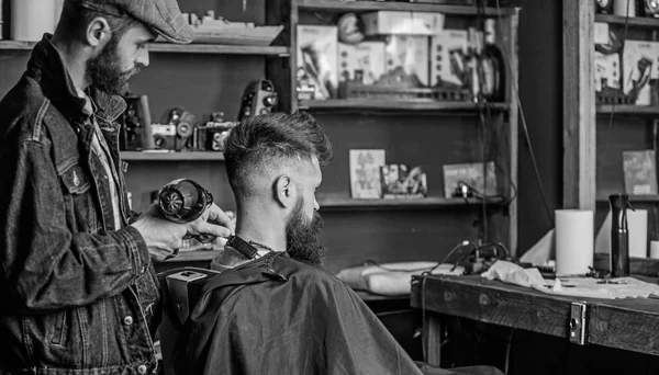 Conceito de barbearia. Hipster barbudo cliente tem penteado. Barbeiro com secador de cabelo funciona no penteado para homem barbudo, barbearia fundo. Barbeiro com secador de cabelo tira o cabelo da capa — Fotografia de Stock