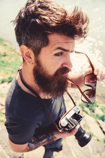 Homme à la barbe et à la moustache met des lunettes de soleil, surface de l'eau sur le fond. Hipster sur le visage strict tient l'appareil photo à l'ancienne. Concept de photographe touristique. Homme tirant sur la nature près de la rivière ou étang — Photo