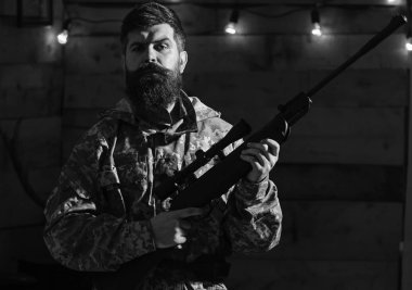 Sakallı adam ahşap iç arka planda kamuflaj elbiseler giyer. Hakem kavramı. Avcı, av için hazır elinde tabanca ile acımasız hipster. Maço gamekeepers evinde sıkı yüzünde