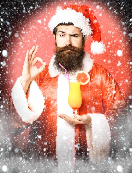 Schöner bärtiger Weihnachtsmann mit langem Bart auf ernstem Gesicht, der ein Glas alkoholfreien Cocktails in Weihnachts- oder Weihnachtspullover und Neujahrsmütze auf rotem Studiohintergrund hält — Stockfoto