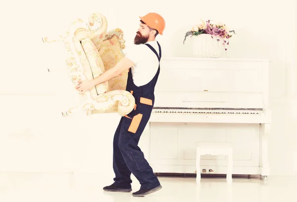 Relocating concept. Man with beard, worker in overalls and helmet lifts up armchair, white background. Courier delivers furniture in case of move out, relocation. Loader carries armchair