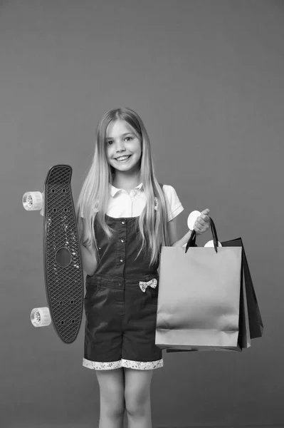 Chica feliz mantenga penny board y bolsas de compras sobre fondo violeta. Sonrisa infantil con paquetes y monopatín. Patinadora sonriendo con bolsas de papel. Compras y venta. Deporte y hobby activo —  Fotos de Stock