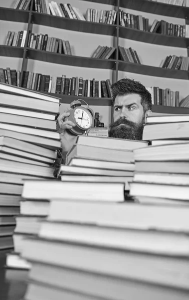 Conceito prazo. Homem de cara dura a olhar para o relógio, prateleiras de livros no fundo. Professor ou estudante com barba estudando na biblioteca. Homem, cientista a espreitar de montes de livros com despertador — Fotografia de Stock