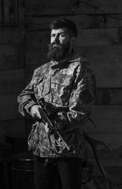 Sakallı kamuflaj elbiseler, ahşap iç arka plan giyer. Avcı, acımasız hipster silahla avlamaya hazır. Avcı kavramı. Maço gamekeepers evinde sıkı yüzünde