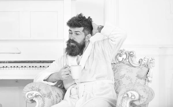 Man with beard and mustache enjoys morning while sitting on old fashioned luxury armchair. Man sleepy in bathrobe drinks coffee in luxury hotel in morning, white background. Morning coffee concept