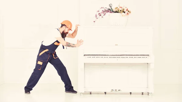Man with beard, worker in overalls and helmet pushes piano, white background. Courier delivers furniture in case of move out, relocation. Delivery service concept. Loader moves piano instrument