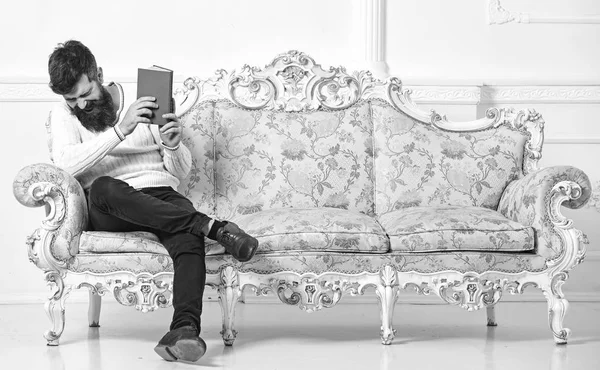 Facet czytając stare książki z przyjemności. Koncepcja literatury humorystyczny. Człowiek z broda i wąsy siedzi na styl barokowy sofa, posiada książkę, tło białe ściany. Macho się ze śmiechu czytając książkę twarz — Zdjęcie stockowe