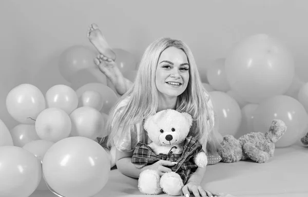 Birthday girl concept. Woman cute celebrate birthday with balloons. Girl in pajama, domestic clothes lay near air balloons, pink background. Blonde on smiling face relaxing with teddy bear toy