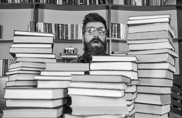 Ο άνθρωπος, nerd στο έκπληκτος πρόσωπο μεταξύ σωρούς από βιβλία στη βιβλιοθήκη, ράφια σε φόντο. Έννοια του σπασίκλα. Δάσκαλος ή μαθητής με γενειάδα φοράει γυαλιά, κάθεται στο τραπέζι με τα βιβλία, defocused — Φωτογραφία Αρχείου
