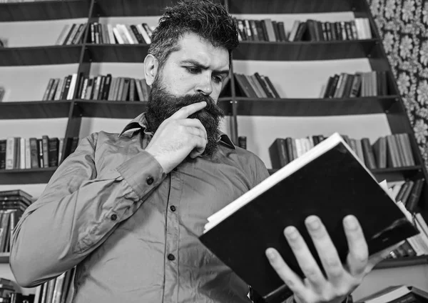 Man on busy thoughtful face reading book, bookshelves on background. Education and science concept. Teacher or student with beard studying in library. Scientist busy with exploring book