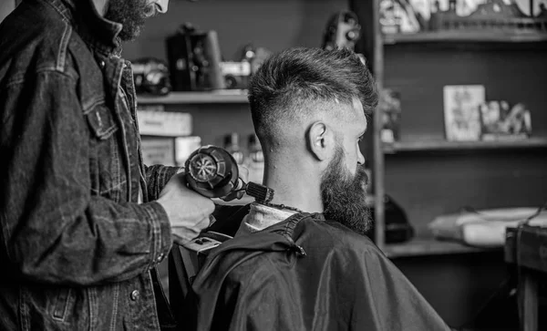 Barbershop concept. Hipster bearded client got hairstyle. Barber with hairdryer blows off hair out of cape. Barber with hairdryer works on hairstyle for bearded man, barbershop background — Stock Photo, Image