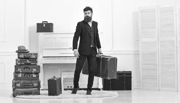 Concepto de equipaje y vacaciones. Macho elegante en la cara estricta se encuentra cerca de la pila de maleta vintage, sostiene la maleta. Hombre, viajero con barba y bigote con equipaje, fondo interior blanco de lujo — Foto de Stock