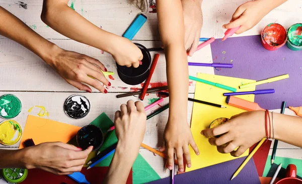 Artists hands with stationery and colored paper. Art supplies in male and female hands on white wooden desk background, top view. Hands hold colorful markers, pencils and paints. Messy art concept