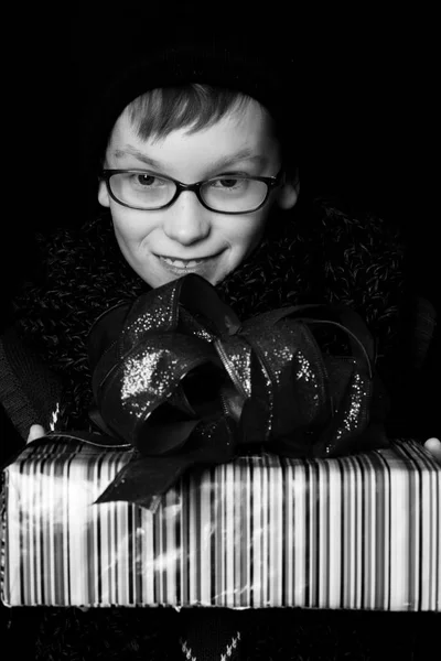 Pequeño niño sonriente o lindo niño nerd en gafas, sombrero y bufanda de punto de moda sobre fondo negro sostiene caja de regalo colorido con bo rojo —  Fotos de Stock