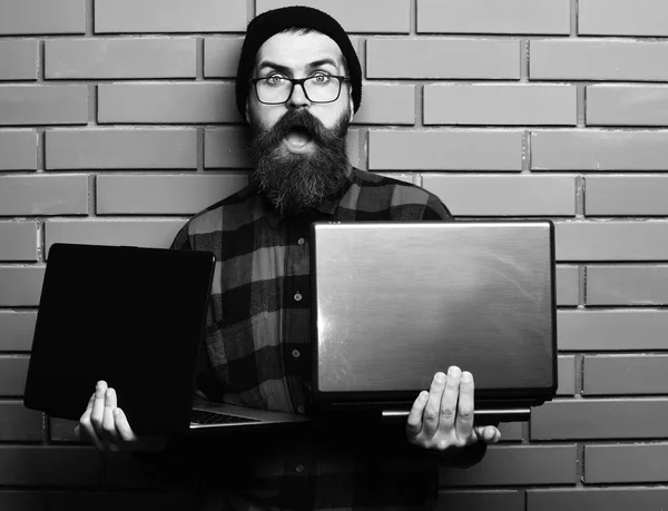 Homem barbudo, barba comprida. Brutal caucasiano surpreendido unshaven hipster segurando laptops em vermelho camisa xadrez preto com chapéu e óculos no backgroun estúdio de parede de tijolo marrom — Fotografia de Stock