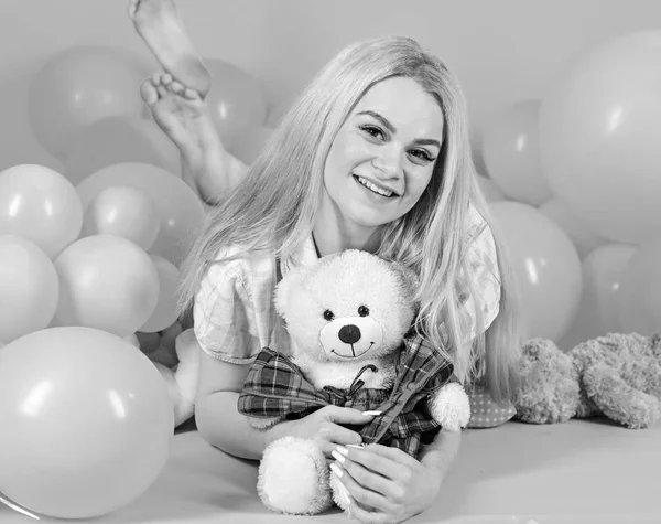 Молода жінка на ліжку обіймає плюшевого ведмедя. Концепція дівчини на день народження. Блондинка на усміхненому обличчі розслабляється іграшкою плюшевого ведмедя. Жінка мило відзначає день народження повітряними кулями. Дівчина в піжамі, рожевий фон — стокове фото