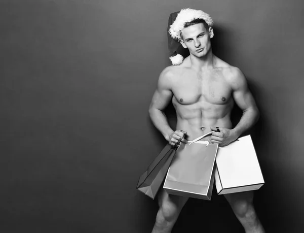 Giovane bell'uomo macho Santa con sexy muscoloso atletico corpo forte ha torso nudo e pancia forte a Natale o Natale cappello tenendo borse regalo colorate su sfondo studio verde, copia spac — Foto Stock