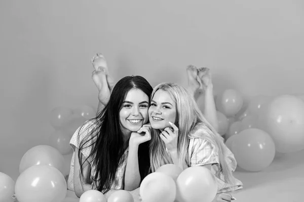 Blonde and brunette on smiling faces have fun at domestic party. Girls lay on belly near balloons, pink background. Friendship concept. Sisters or best friends in pajamas at girlish pajamas party — Stock Photo, Image