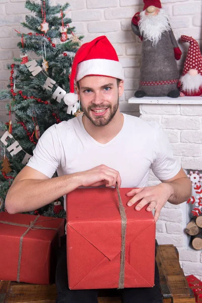 Man in santa hat hold christmas present. delivery christmas gifts. online christmas shopping. New year scene with tree and gifts. happy santa man. The morning before Xmas. Office christmas party — Stock Photo, Image