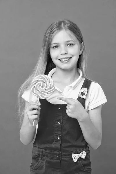 Menina alegre bonito segurando doces gigantes. Criança com cabelo loiro longo isolado em fundo violeta. Criança apontando para pirulito colorido, conceito de infância doce. Menina em macacão rosa gosta de doces . — Fotografia de Stock