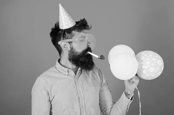 Homem com barba aparada usando camisa rosa e boné de festa isolado no fundo vermelho. Homem barbudo soprando assobiar festa e segurando balões brilhantes. Hipster com barba longa em óculos enormes, conceito divertido — Fotografia de Stock