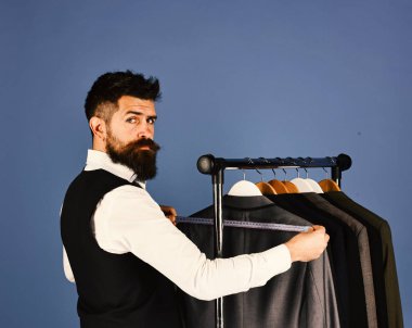 Yelekli sakallı adam elbise rafında. Tasarımcı elbise askılarının yanında önlem alır. Moda seçimi konsepti. Ciddi yüzlü terzi, mavi arka plandaki özel ceketlerin yanında mezura tutuyor..