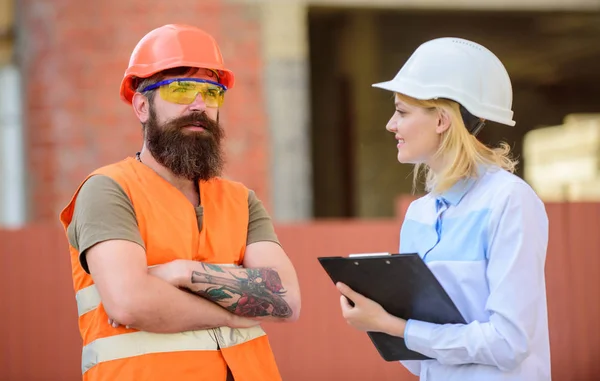 Woman inspector and bearded brutal builder discuss construction progress. Construction project inspecting. Construction site safety inspection. Discuss progress project. Safety inspector concept