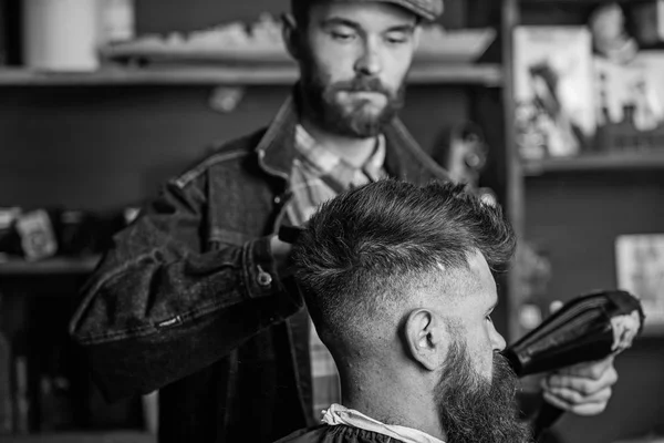 Hipster bearded client getting hairstyle. Styling concept. Barber with hairdryer drying and styling hair of client. Barber with hairdryer works on hairstyle for bearded man, barbershop background