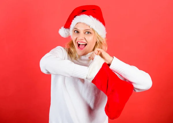 Mädchen fröhliches Gesicht check out Geschenk in Weihnachtssocke. Frau mit Weihnachtsmütze auspacken Weihnachtsgeschenk roten Hintergrund. Kontrollieren Sie den Inhalt von Weihnachtsstrumpf, was sie erhalten hat. Weihnachtsstrumpfkonzept — Stockfoto