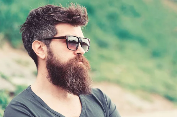 Hipster man in trendy sunglasses standing on slope with lush green glass in spring. Bearded man with stylish hairstyle enjoying the view over city