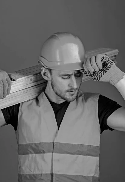 Carpenter, woodworker, strong builder on busy face carries wooden beam on shoulder. Safety and protection concept. Man in protective gloves holds visor of protective hard hat, grey background — Stock Photo, Image