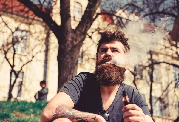 Vaping en gewoonten concept. Man met baard en snor roken, stedelijke achtergrond. Hipster met verwarde haren en grijs op ontspannen dromerige gezicht met witte rook uit mond vliegen — Stockfoto