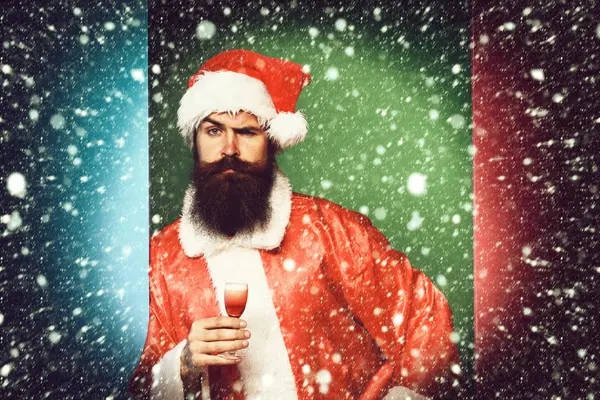 Guapo barbudo hombre de Santa Claus con barba larga en la cara seria sosteniendo vaso de trago alcohólico en navidad roja o suéter de Navidad y sombrero de año nuevo en fondo colorido estudio —  Fotos de Stock