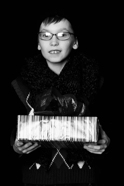 Pequeño niño sonriente o lindo niño nerd en gafas, sombrero y bufanda de punto de moda sobre fondo negro sostiene caja de regalo colorido con bo rojo — Foto de Stock