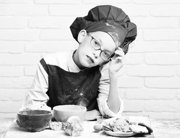 Young boy cute cook chef in red uniform and hat on stained face with glasses sitting on table with colorful bowls, tasty cakes, rolling pin and kitchen whisk on white brick wall backgroun — Stock Photo, Image